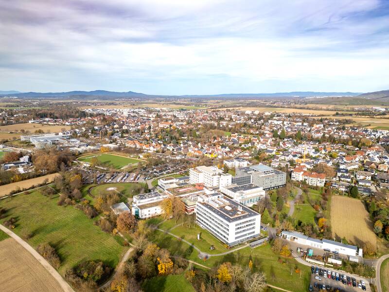 Eine weite Landschaft mit Feldern in unterschiedlichen Grüntönen erstreckt sich vor einer Bad Krozingen im Hintergrund, eingerahmt von Hügeln des Schwarzwald unter einem freundlichem Himmel.