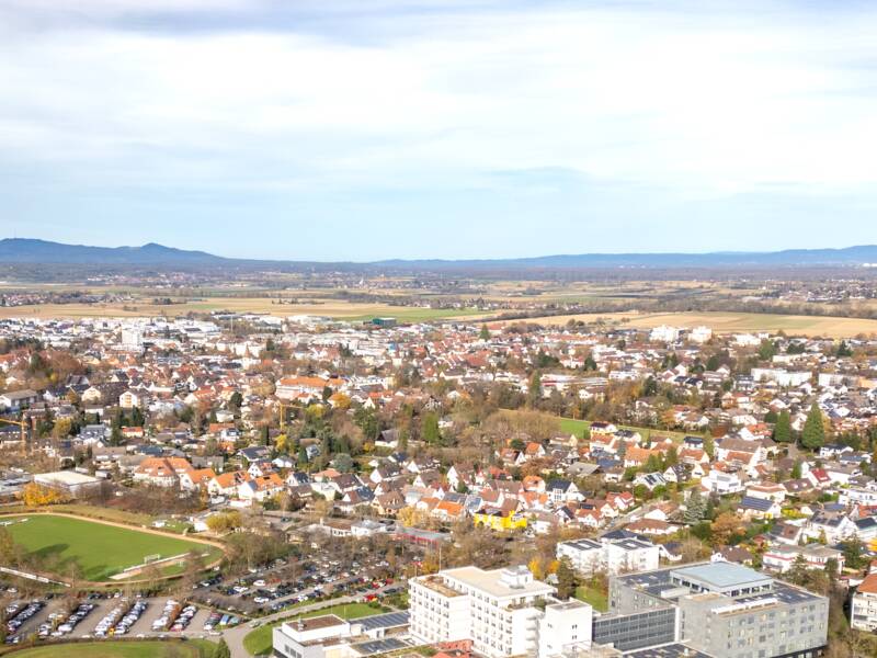 Eine weite Landschaft mit Feldern in unterschiedlichen Grüntönen erstreckt sich vor einer Bad Krozingen im Hintergrund, eingerahmt von Hügeln des Schwarzwald unter einem freundlichem Himmel.