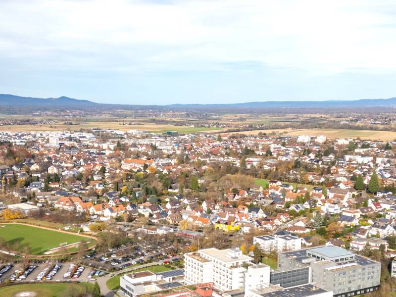 Eine weite Landschaft mit Feldern in unterschiedlichen Grüntönen erstreckt sich vor einer Bad Krozingen im Hintergrund, eingerahmt von Hügeln des Schwarzwald unter einem freundlichem Himmel.