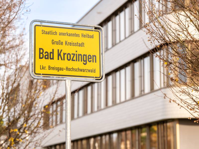 Ein gelbes Ortsschild mit der Aufschrift "Bad Krozingen" steht vor einem modernen Gebäude mit Bäumen im Vordergrund.