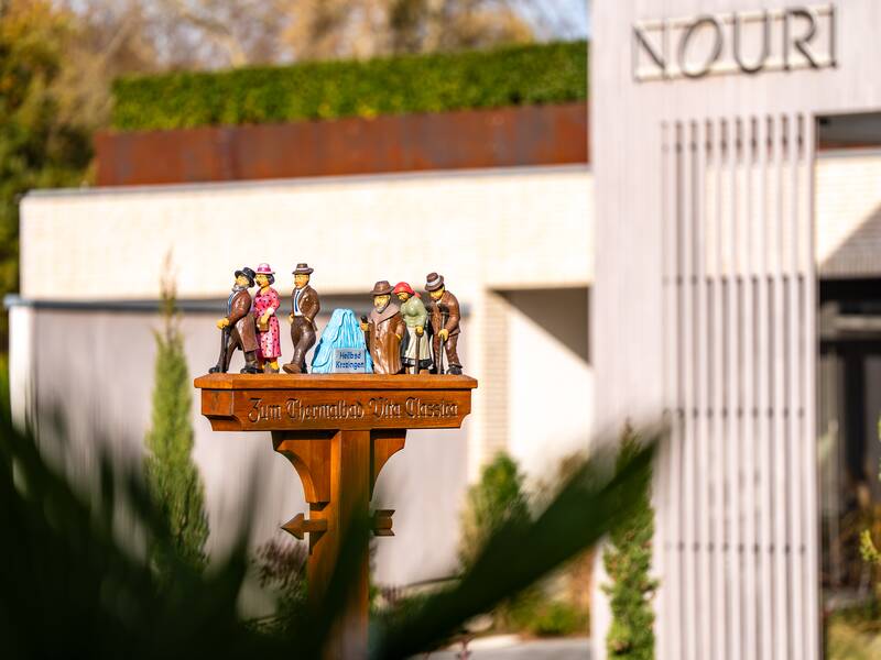 Eine Holzskulpturengruppe, die mehrere Figuren in traditioneller Kleidung darstellt, steht auf einem hölzernen Podest mit der Inschrift "Zum Thermalbad Vita Classica".