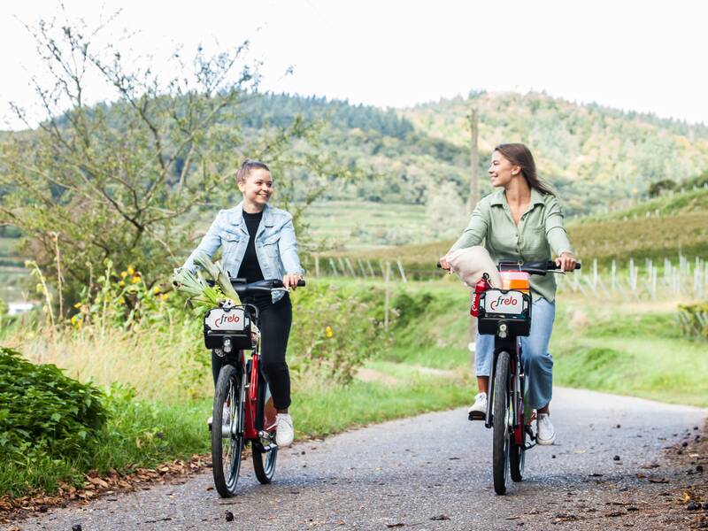 Zwei Frauen fahren, jeweils mit Taschen in Fahrradkörben, fröhlich auf einem ruhigen, ländlichen Weg Fahrrad.