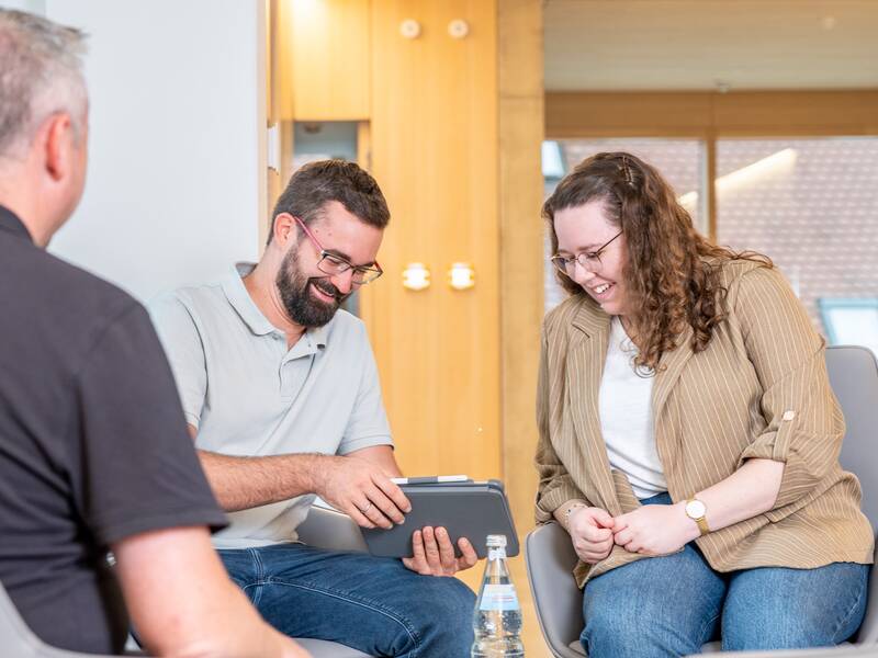 Drei Personen sitzen in einem hellen Raum zusammen und schauen lächelnd auf ein Tablet.