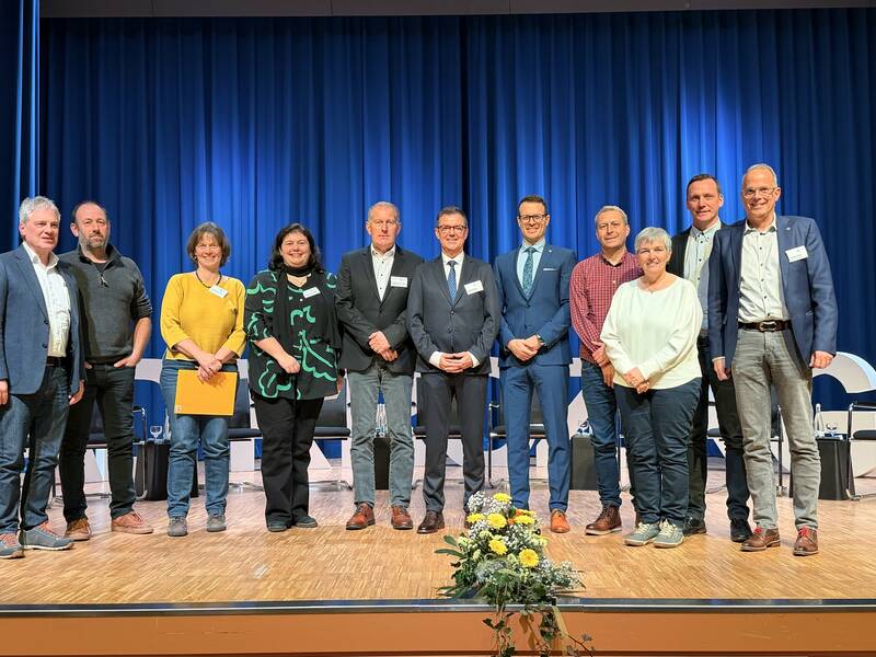 Eine Gruppe von elf Personen steht lächelnd in einer Reihe auf einer Bühne vor einem blauen Vorhang.