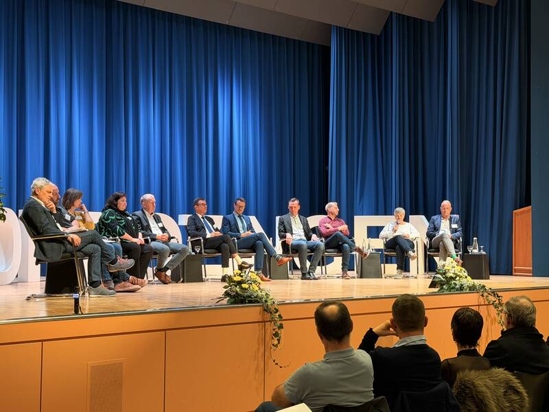 Eine Gruppe von Menschen sitzt auf einer Bühne mit blauen Vorhängen, während sie Teil einer Podiumsdiskussion oder Veranstaltung sind.