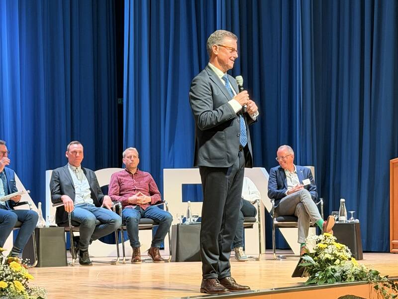 Oberbürgermeister Kieber steht auf einer Bühne und spricht in ein Mikrofon, während mehrere Personen hinter ihm auf Stühlen sitzen und ihm zuhören.