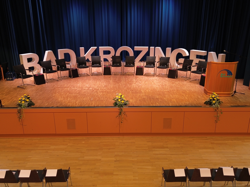 Eine Bühne mit Stühlen, einem Rednerpult und großen weißen Buchstaben, die "BAD KROZINGEN" buchstabieren, ist mit blauen Vorhängen im Hintergrund dekoriert.