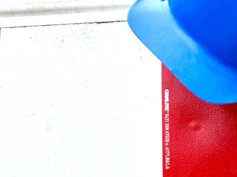 Ein blauer Bauhelm hängt an einer rot-weißen Absperrtafel mit einem blinkenden roten Warnlicht darauf, vor einem grünen Baum im Hintergrund.