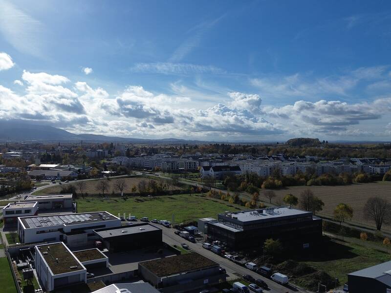Eine Luftaufnahme zeigt eine urbane Landschaft mit Gebäuden, Grünflächen und Feldern unter einem bewölkten Himmel.