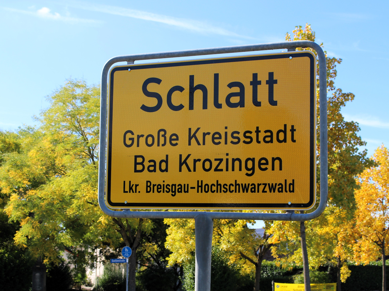 Ein gelbes Ortsschild mit der Aufschrift "Schlatt, Große Kreisstadt Bad Krozingen, Lkr. Breisgau-Hochschwarzwald" steht vor einem Hintergrund aus grünen und gelben Bäumen.