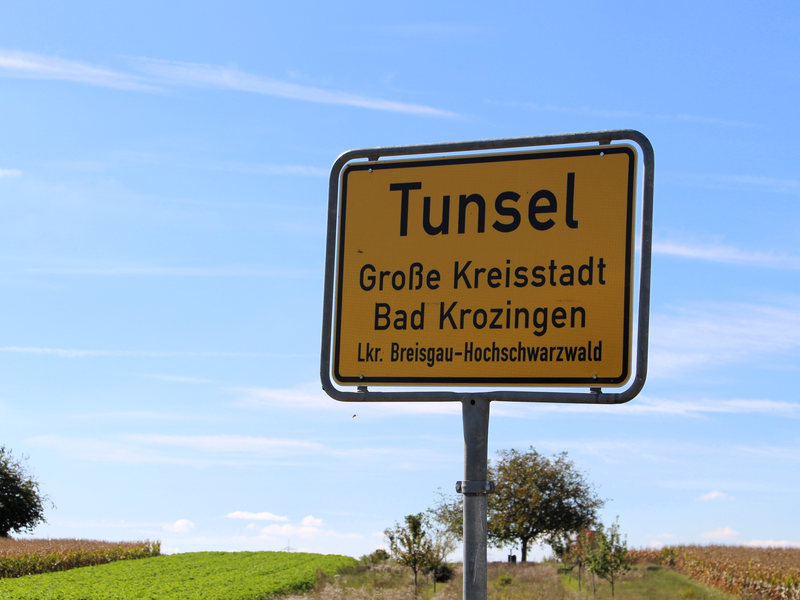 Ein gelbes Ortsschild mit der Aufschrift "Tunsel, Große Kreisstadt Bad Krozingen, Lkr. Breisgau-Hochschwarzwald" steht vor einem Hintergrund aus grünen und gelben Feldern.