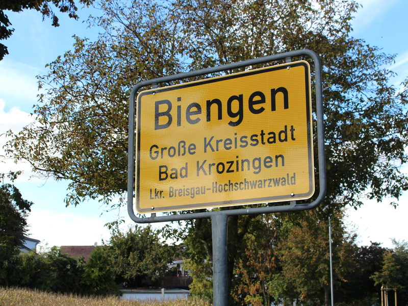Ein gelbes Ortsschild mit der Aufschrift "Biengen, Große Kreisstadt Bad Krozingen, Lkr. Breisgau-Hochschwarzwald" steht vor einem Hintergrund aus grünen Bäumen.