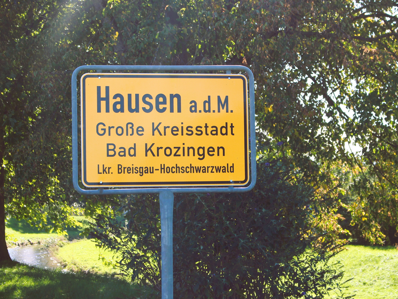 Ein gelbes Ortsschild mit der Aufschrift "Hausen, Große Kreisstadt Bad Krozingen, Lkr. Breisgau-Hochschwarzwald" steht vor einem Hintergrund aus grünen Bäumen und dem Fluss Möhlin.