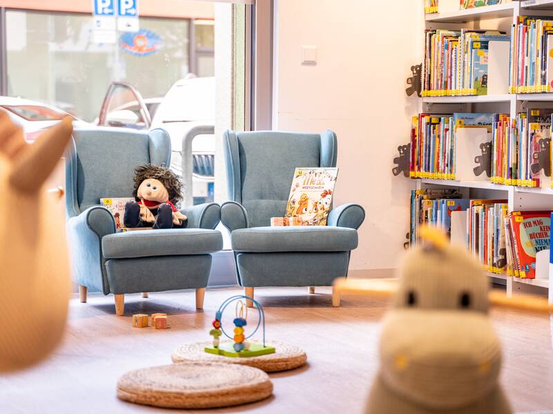 Eine gemütliche Leseecke mit zwei blauen Sesseln, auf denen Puppen und Kinderbücher sitzen, umgeben von Bücherregalen, lädt zum Verweilen ein.