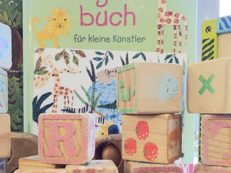 Mehrere bunte Holzbausteine mit Buchstaben und Zahlen sind vor einer Sammlung von Kinderbüchern auf einem Tisch gestapelt.