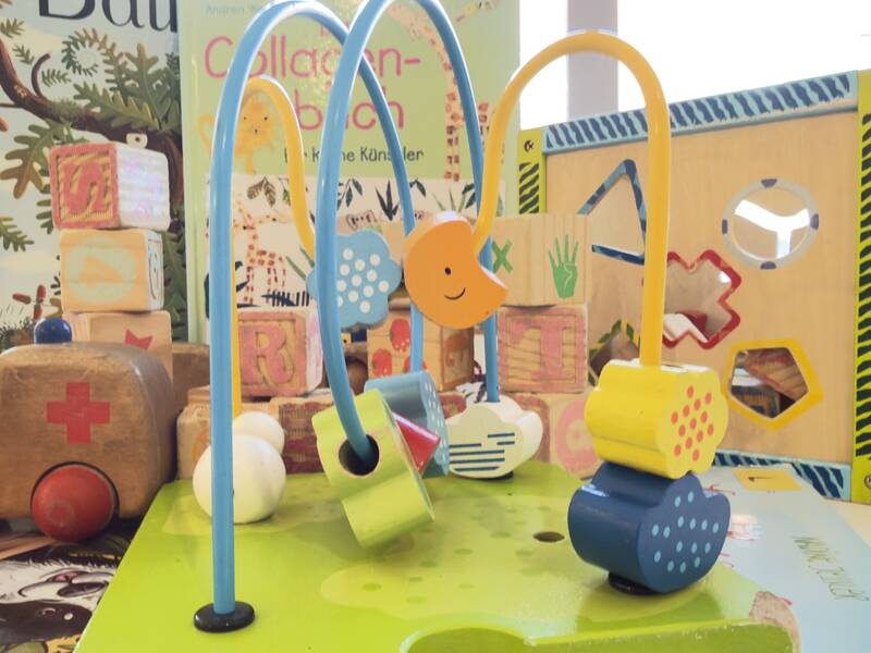 Auf dem Bild sind verschiedene bunte Kinderspielzeuge aus Holz, darunter ein Spielzeugauto, Bausteine, eine Motorikschleife und ein Formen-Steckwürfel, vor einigen Kinderbüchern zu sehen.