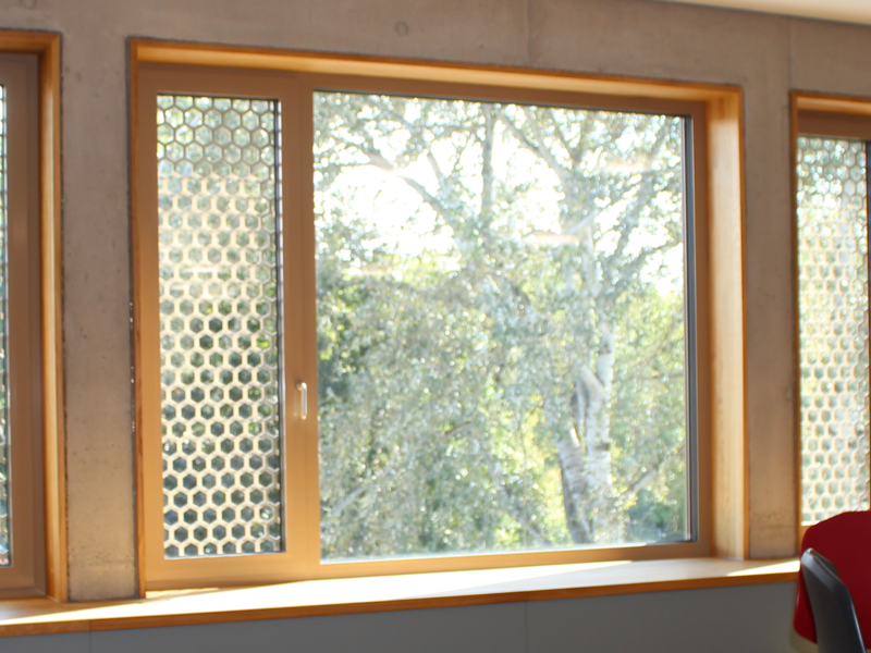 Der Raum verfügt über große Fenster mit wabenförmigen Gittern, eine gebogene Sitzbank aus Holz und moderne, rot-schwarze Stühle an runden Tischen.