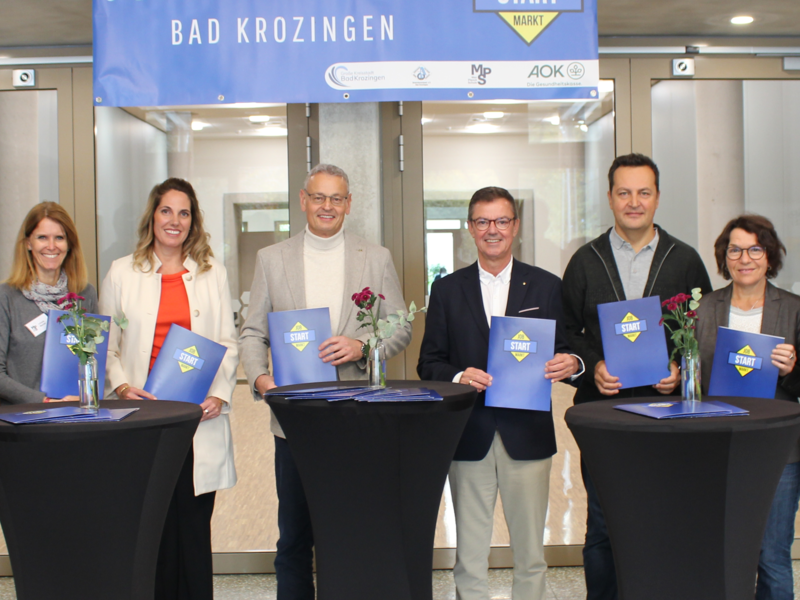 Eine Gruppe von neun Personen steht vor einem Banner mit der Aufschrift "Jobstartmarkt Bad Krozingen" und hält blaue Broschüren in den Händen.