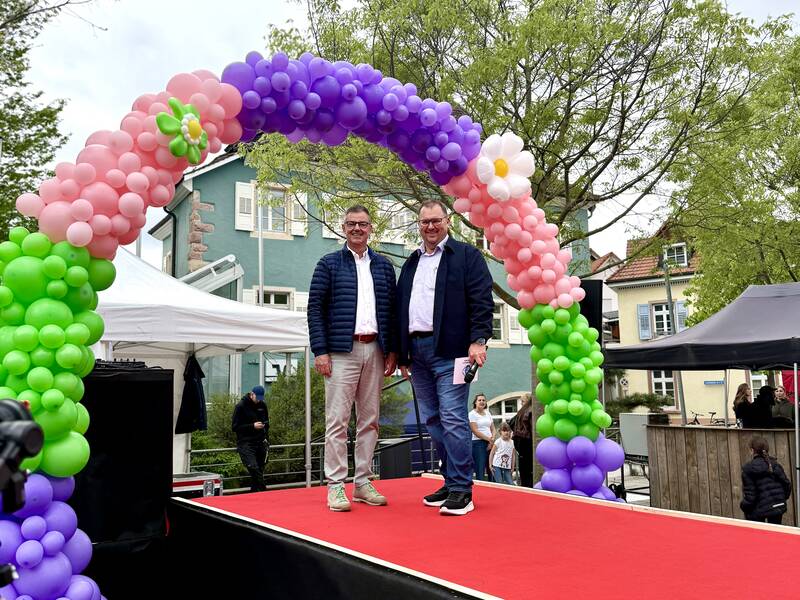 Zwei Personen, Oberbürgermeister Volker Kieber und Gewerbevorstand Peter Lob stehen auf einer roten Plattform unter einem farbenfrohen Torbogen aus rosa, lila und grünen Luftballons, während im Hintergrund ein Gebäude und Bäume zu sehen sind.