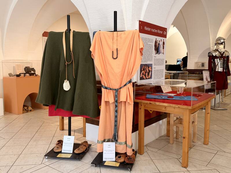 In einem Ausstellungsraum sind historische Römerkleidungsstücke und Rüstungen aufgestellt, begleitet von Informationstafeln.