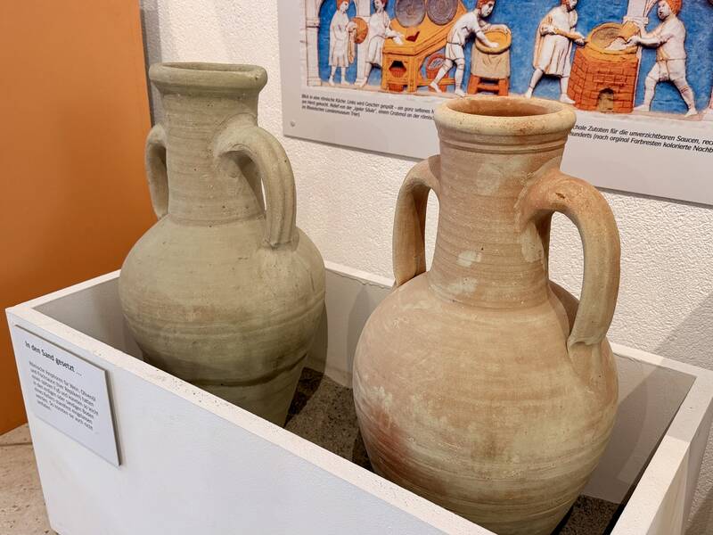 Zwei große, beige Tonkrüge mit je zwei Henkeln stehen in einem Museumsdisplay, daneben befindet sich ein informatives Poster mit einer Illustration im Hintergrund.