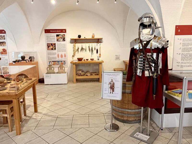 Das Bild zeigt eine Museumsausstellung mit römischen Artefakten, einem römischen Kostüm und Infotafeln in einem Raum mit gewölbter Decke.