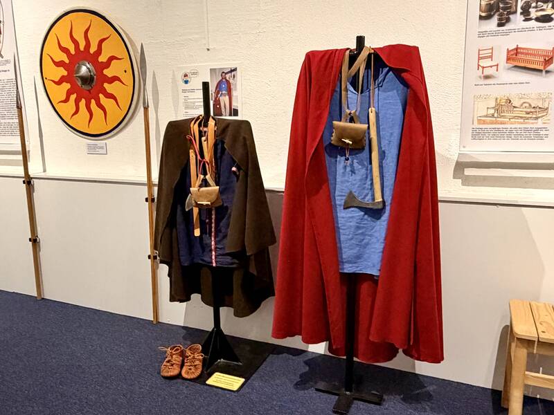In einem Ausstellungsraum sind historische Kleidungsstücke, ein gelber Rundschild mit rotem Sonnenmotiv und Speere ausgestellt.