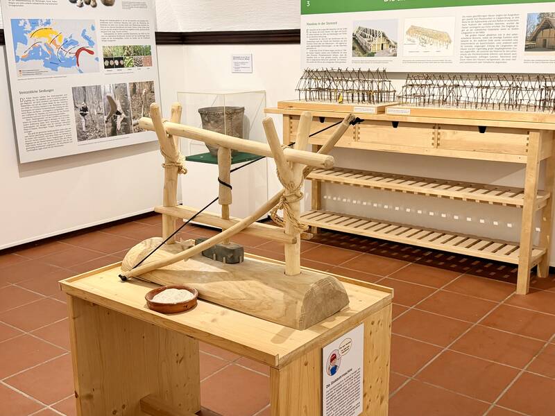 Ein Ausstellungsraum zeigt ein hölzernes Modell einer historischen Konstruktion auf einem Holztisch, mit erklärenden Postern und Modellen im Hintergrund.