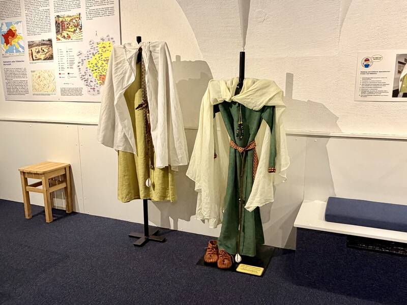 In einem Ausstellungsraum sind zwei historische Gewänder auf schwarzen Ständern zu sehen, umgeben von erklärenden Texttafeln an der Wand.