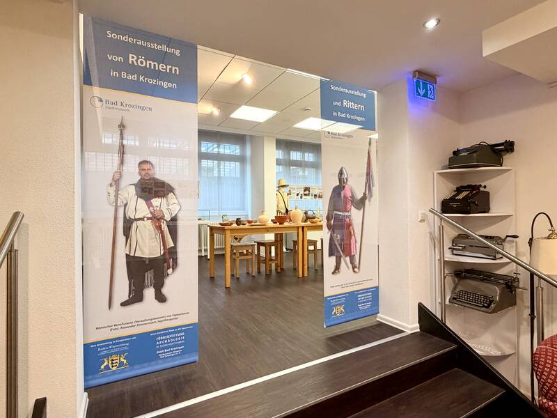 Ein Ausstellungsraum mit Bannern, die auf eine Sonderausstellung über Römer und Ritter in Bad Krozingen hinweisen, eingerahmt von historischen Schreibmaschinen und einem Lesestuhl.