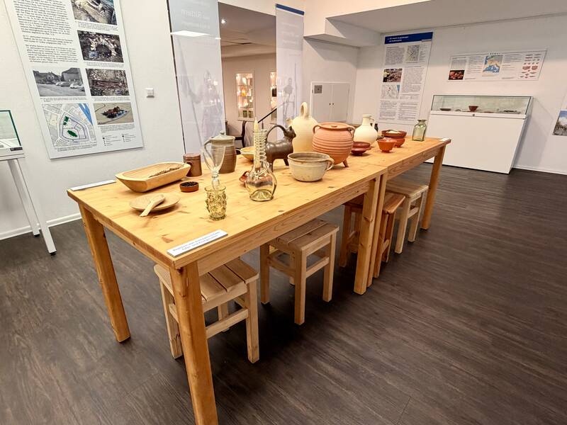Ein Ausstellungsraum zeigt einen langen Holztisch mit verschiedenen Tongefäßen und anderen Gegenständen darauf, umgeben von Infotafeln an den Wänden.