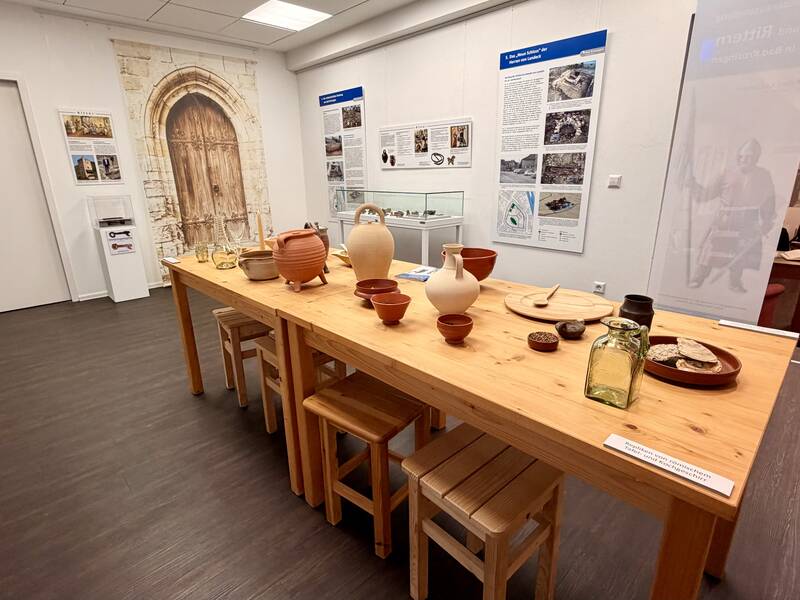 Ein moderner Ausstellungsraum mit einem langen Holztisch in der Mitte, auf dem antike Gefäße und Nachbildungen antiker Gegenstände stehen, umgeben von Informationstafeln an den Wänden.