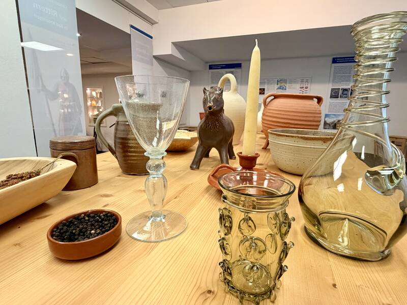 Eine Ausstellung von antiken Haushaltsgegenständen und Geschirr, darunter Gläser, Keramik, eine Kerze und Behälter aus Ton und Glas, auf einem Holztisch in einem Museum.