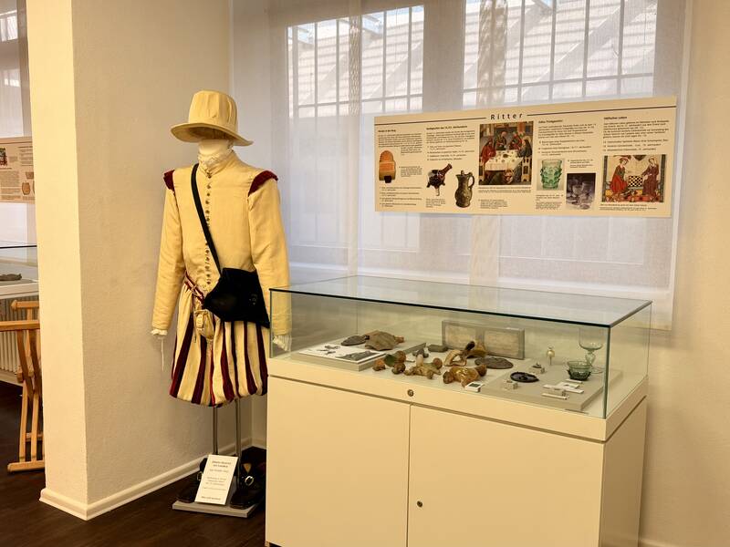 Ein Ausstellungsraum in einem Museum zeigt eine historische Figur in mittelalterlicher Kleidung neben einer Vitrine mit Artefakten und Informtaionstafeln an den Wänden.