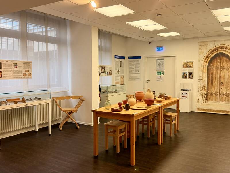 Ein Raum mit Ausstellungsstücken und Archäologie-Objekten in Glasvitrinen und auf einem Holztisch, umgeben von Informationstafeln und einem großen Türbild an der Wand.