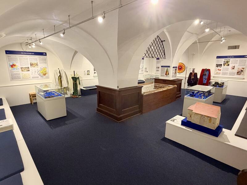 Ein Ausstellungsraum mit weißen Gewölbedecken zeigt historische Kleidung, Exponate in Vitrinen und Informationstafeln an den Wänden.