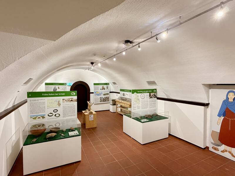 Ein heller Museumsraum mit Vitrinen und Informationstafeln zu Ausgrabungsfunden sowie einer Abbildung einer Person an der Wand.