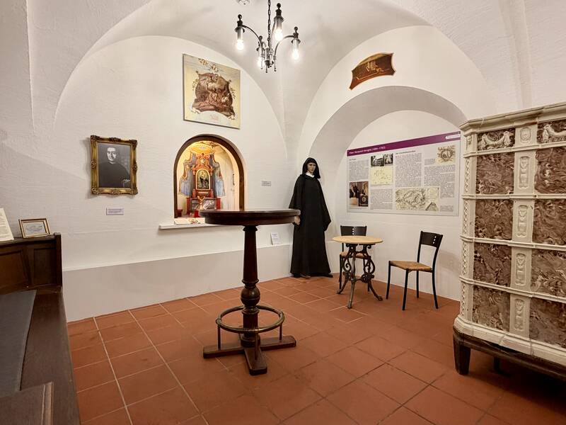 Ein Ausstellungsraum mit historischen Gegenständen, einem alten Ofen, einem runden Tisch, einem Porträt und einer bekleideten Schaufensterpuppe vor illustrierten Tafeln und Bildern.