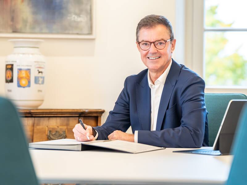 Oberbürgermeister Kieber sitzt in einem Anzug lächelnd an einem Tisch und unterschreibt Dokumente.