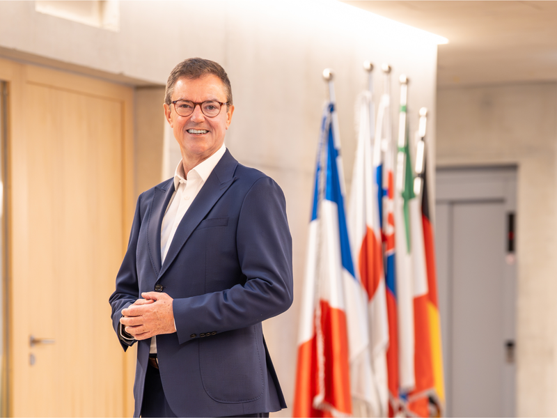 Oberbürgermeister Kieber steht  lächelnd in einem Flur vor einer Reihe von europäischen Nationalflaggen.