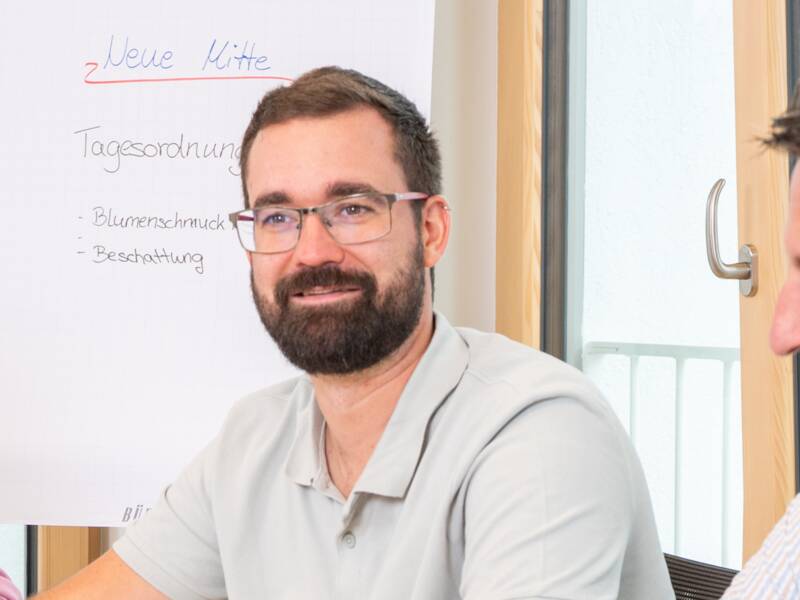 Fünf Personen sitzen an einem Tisch in einem Büro und besprechen etwas, während einige von ihnen Laptops oder Tablets benutzen.