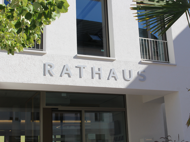 Das Bild zeigt die Fassade eines Gebäudes mit dem Wort "Rathaus" deutlich erkennbar über dem Eingangsbereich, umgeben von einigen Pflanzen.