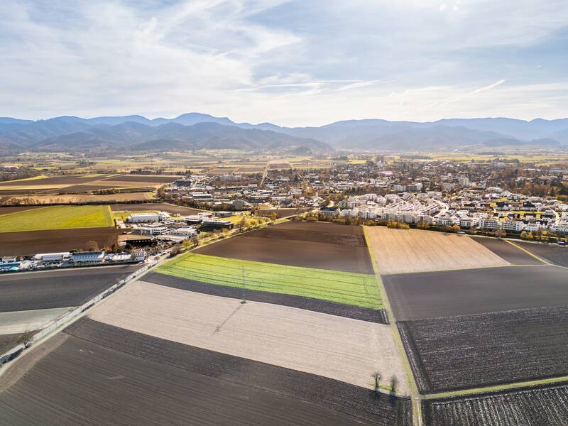 Eine Luftaufnahme zeigt ein weitläufiges, landwirtschaftlich genutztes Gebiet mit Feldern und einer Siedlung im Hintergrund, umgeben von Hügeln und Bergen.