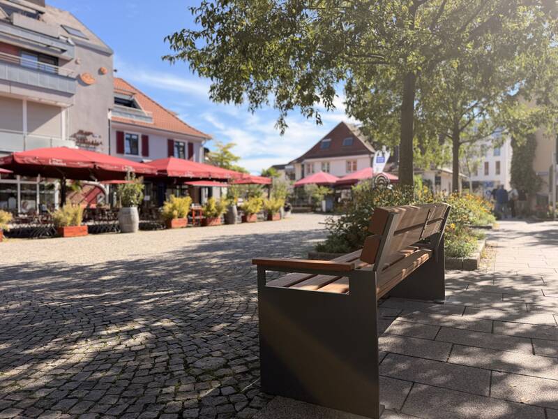 Eine sonnige Straßenszene mit einer Holzbank im Vordergrund und einem Café mit roten Sonnenschirmen umgeben von Pflanzen im Hintergrund.