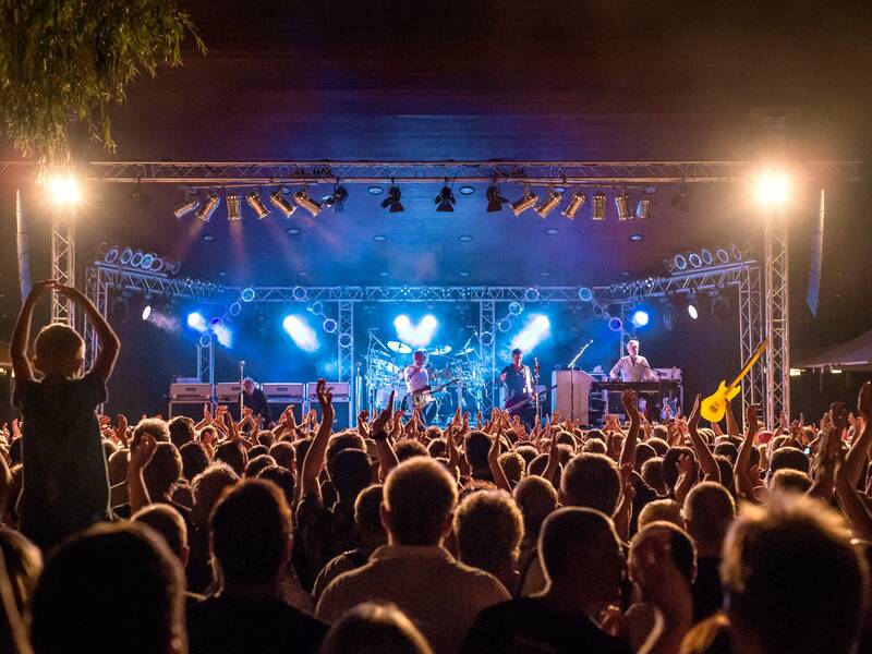 Eine große Menschenmenge steht vor einer beleuchteten Bühne, auf der eine Band spielt, während die Zuschauer jubeln und applaudieren.