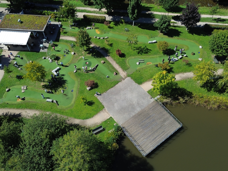 Die Luftaufnahme zeigt eine Mini-Golf-Anlage in einer grünen Parklandschaft mit Gehwegen und einem angrenzenden Teich.