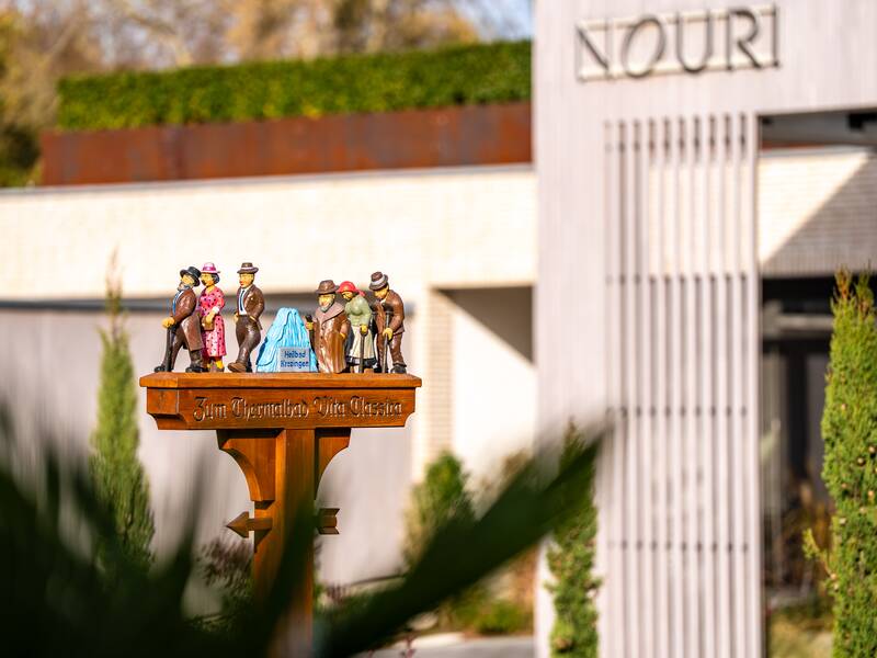 Ein kunstvoll gestalteter Wegweiser mit kleinen, bunten Figuren steht vor einem modernen Gebäude mit der Aufschrift "NOURI".