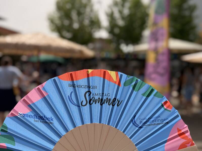 Eine Hand hält einen bunten Fächer mit der Aufschrift "Bad Krozinger Sommer" vor einem unscharfen Hintergrund mit Bäumen und Gebäuden.