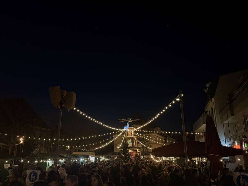 Eine nächtliche Szene eines belebten Weihnachtsmarktes mit festlichen Lichterketten und geschmückten Ständen.
