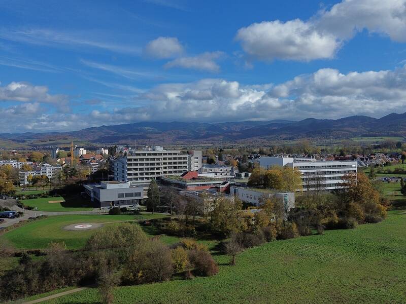 Luftaufnahme einer großen Gebäudestruktur, umgeben von grünen Feldern und Bäumen, mit Bergen im Hintergrund unter einem bewölkten Himmel.